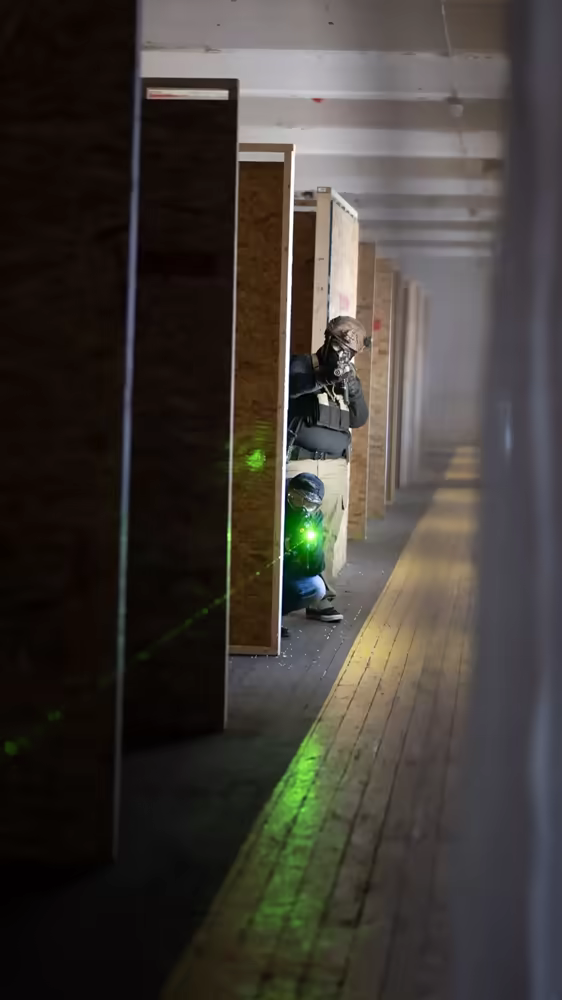 Portrait view of indoor arena corridor with player aiming around a corner, laser beams visible across the room